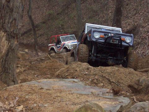 1978 Ford Bronco - Two off-road vehicles tackle a challenging muddy terrain on a wooded trail.