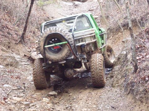 1978 Ford Bronco - A heavily modified green off-road vehicle navigates a steep, muddy incline on a wooded trail.