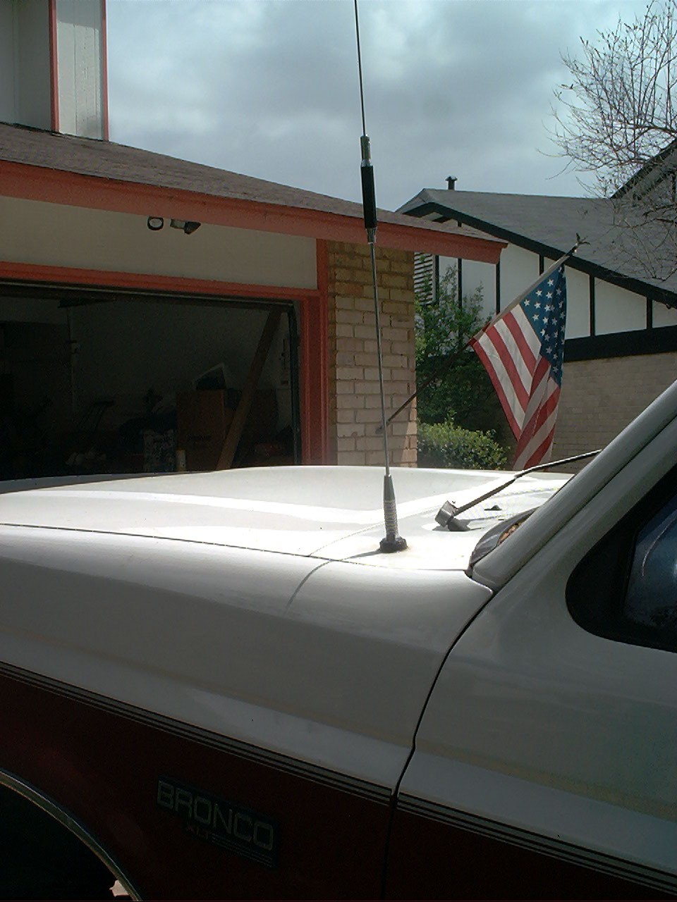 White and red Ford Bronco hood with whip antenna, American flag and house with open garage behind