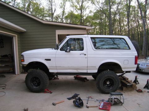 1996 Ford Bronco - A white Ford Bronco is elevated on jack stands, undergoing maintenance in a driveway.