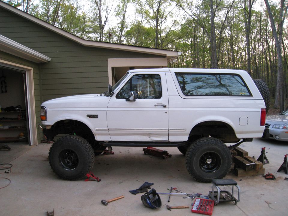 White Ford Bronco on jack stands with large tires, parked in a driveway next to a garage