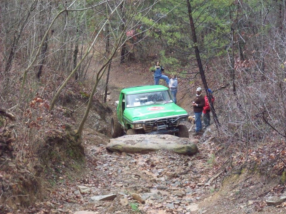 Green off-road truck navigating a rocky, wooded trail with people watching