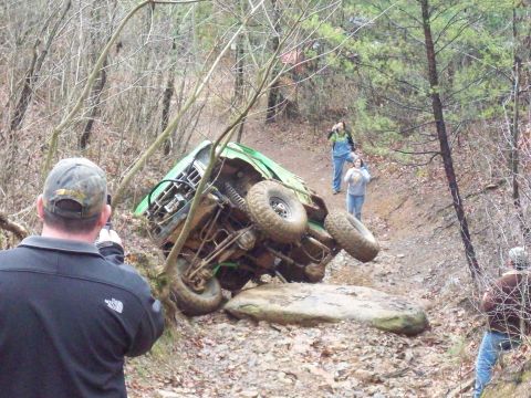 1978 Ford Bronco - Off-road vehicle has tipped over on a rocky, wooded trail, with onlookers observing.