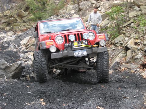 1991 Ford Bronco - A red Jeep Wrangler navigates a challenging off-road course with a person standing nearby.