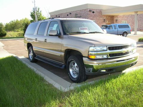 1995 Ford Bronco - A tan Chevrolet Suburban is parked in a lot, with a blue pickup truck visible behind it.