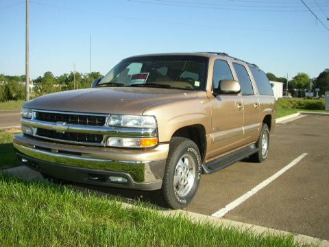 1995 Ford Bronco - A gold Chevrolet Suburban is parked in a lot on a sunny day. A 'For Sale' sign is visible on the windshield.