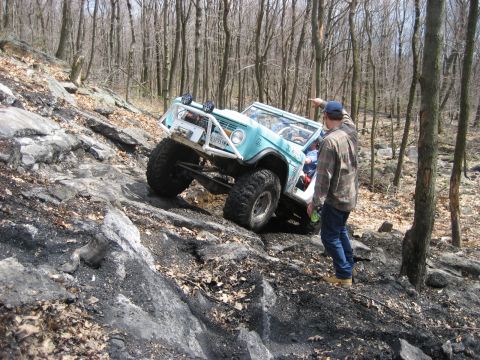 1991 Ford Bronco - A man points while a modified SUV navigates a challenging rocky trail in a wooded area.