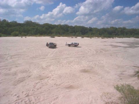 1994 Ford Bronco - Several vehicles are parked in a sandy, open area with trees lining the horizon under a cloudy sky.