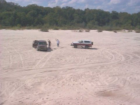 1994 Ford Bronco - Two vehicles are parked in a sandy area with trees in the background.