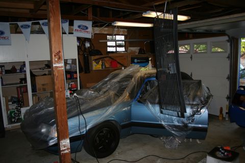 1990 Ford Mustang - Car covered in plastic wrap in a garage, with a grille hanging nearby.