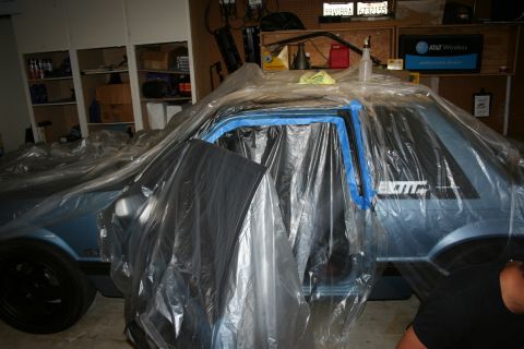 1990 Ford Mustang - A light blue Ford Mustang is covered in plastic and tape, likely being prepped for painting or detailing in a garage.