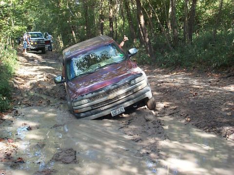 1994 Ford Bronco - A maroon SUV is stuck deep in mud on a wooded trail, with another vehicle visible in the background.