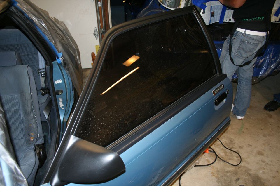Blue 1990 Ford Mustang door open, showing interior and exterior. Tinted window reflects light