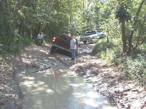 1994 Ford Bronco - Two trucks navigate a challenging muddy path through a wooded area, with one vehicle partially submerged in water.