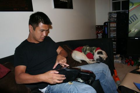 1998 Ford Crown Victoria - man sits on a dark couch holding an object while a pug wearing a camo shirt sleeps beside him.