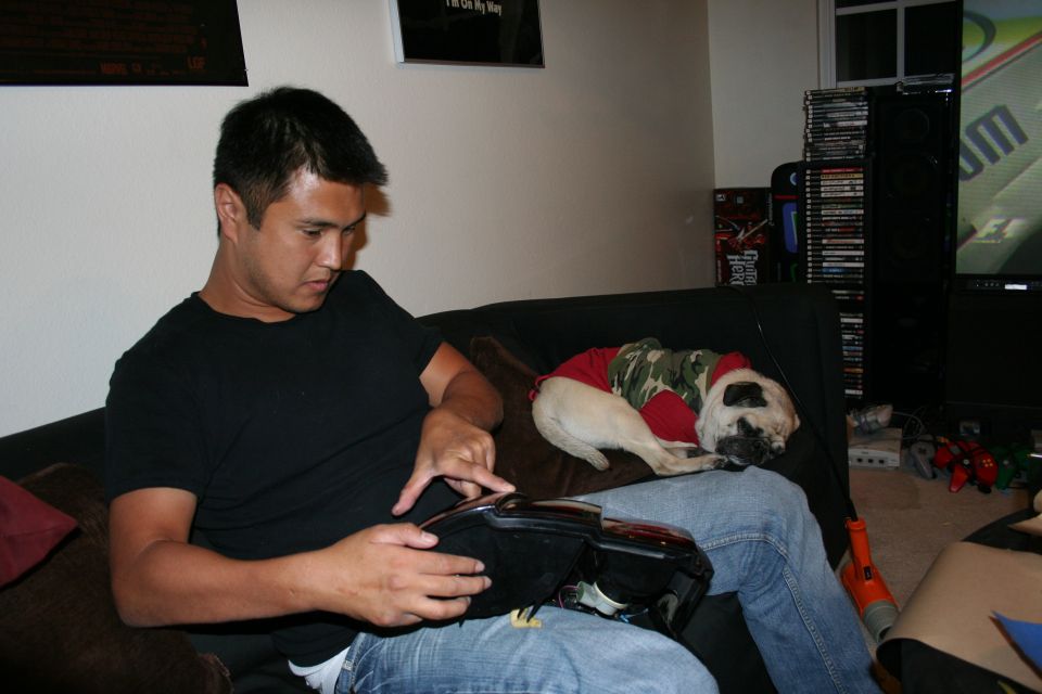 Man in black shirt sitting on a couch beside a sleeping pug in a green shirt