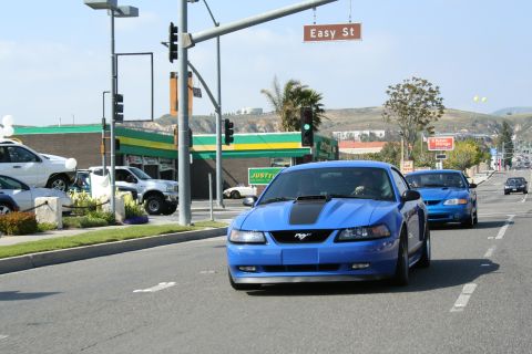 1998 Ford Crown Victoria - blue Ford Mustang with a hood scoop drives on a street under an Easy St sign, a second blue car behind.