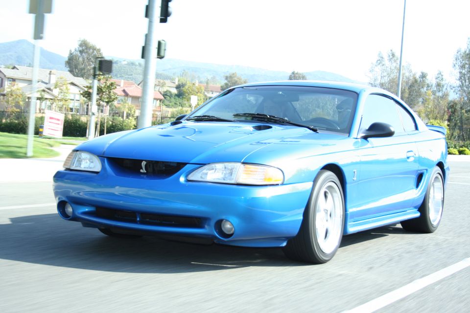Blue Mustang SVT Cobra coupe with white wheels driving on a road