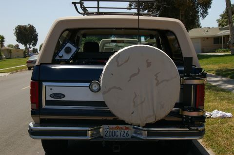 1986 Ford Bronco - The rear of a Ford Bronco is shown, a spare tire cover and a roof rack, parked on a suburban street.
