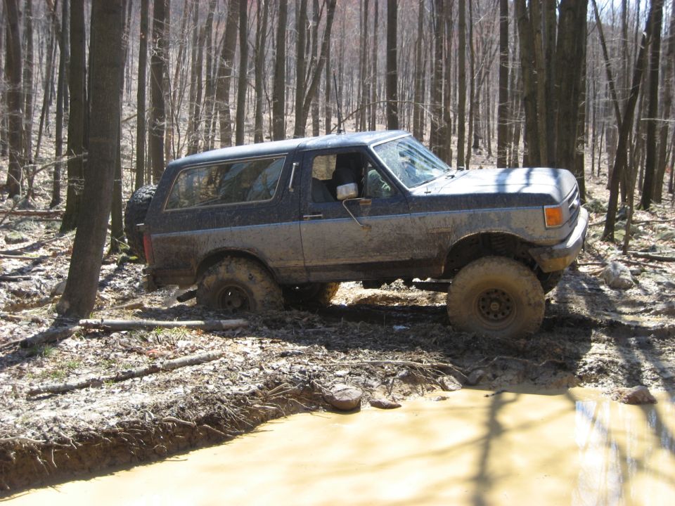 Muddy Ford Bronco navigating a wooded trail with a spare tire on the back