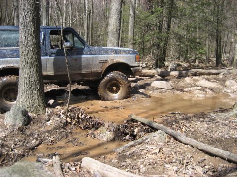 1991 Ford Bronco - A Ford Bronco navigates a challenging muddy path in a wooded area.