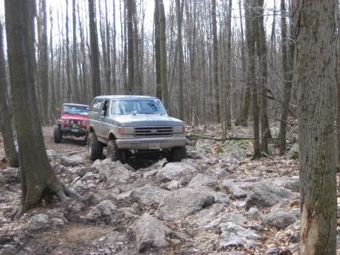 1991 Ford Bronco - A gray 1991 Ford Bronco navigates a challenging rocky trail in a wooded area, followed by a red Jeep.
