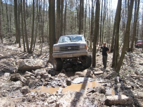 1991 Ford Bronco - A lifted 1991 Ford Bronco tackles a challenging muddy obstacle course in a wooded area, with another vehicle visible in the background.