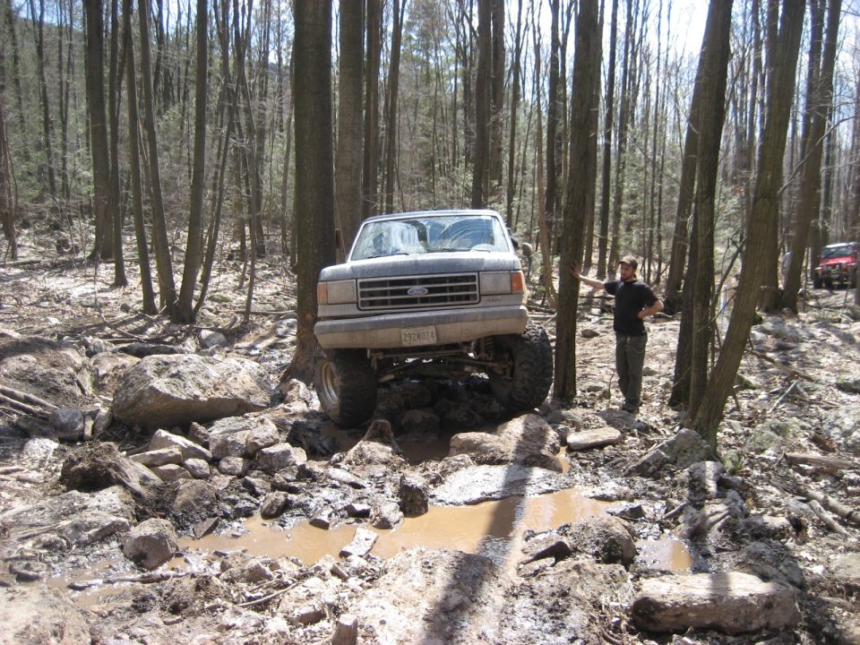 Gray 1991 Ford Bronco navigating a muddy, wooded trail with a person standing nearby