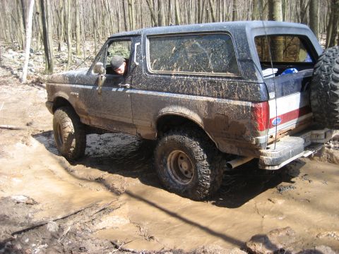 1991 Ford Bronco - A muddy Ford Bronco drives through a wooded area with a spare tire mounted on the back.