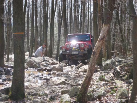 1991 Ford Bronco - A red Jeep Wrangler tackles a challenging off-road course through a dense forest, with a person observing from behind the vehicle.