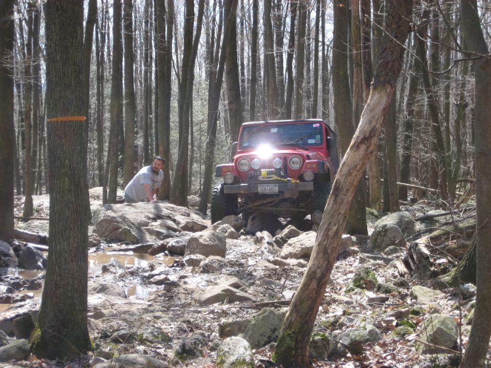 Red Jeep Wrangler navigating a rocky, wooded trail with a person behind it