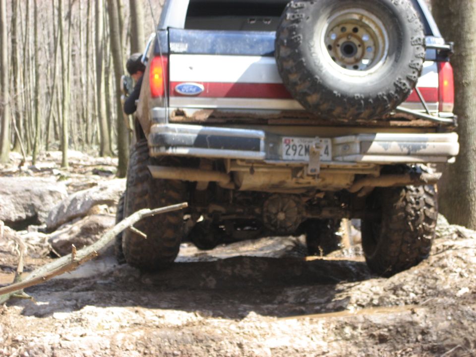 Rear view of a lifted Ford Bronco driving over rocks and mud in a wooded area. It has a spare tire mounted on the back
