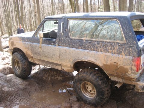 1991 Ford Bronco - A heavily mud-splattered Ford Bronco navigates a challenging off-road course through a wooded area.