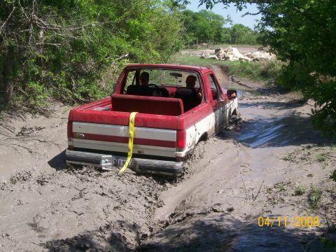 1988 Ford Bronco - This truck is deep in the mud on a trail with trees on either side.