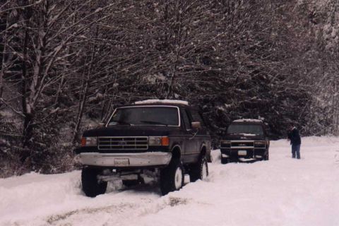 1988 Ford Bronco - Two SUVs are parked on a snow-covered road surrounded by trees.