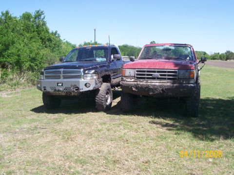 1988 Ford Bronco - Two trucks covered in mud sit side-by-side on a grassy field under a clear blue sky.