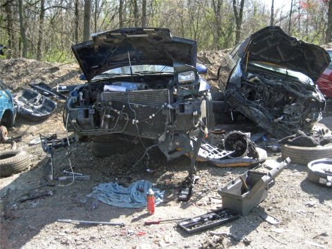 1993 Ford Probe - Junkyard scene with two heavily damaged vehicles and scattered parts under trees.