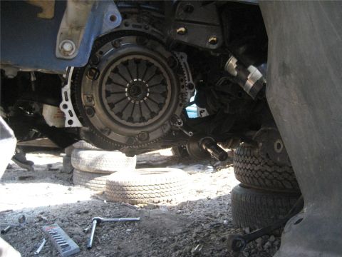 1993 Ford Probe - Vehicle's clutch assembly and transmission are visible, supported by stacked tires, with tools scattered on the dirt ground.