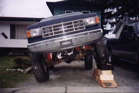 1988 Ford Bronco - This lifted Ford truck features a prominent grille and is parked next to another dark vehicle.