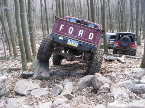 1991 Ford Bronco - A maroon Ford pickup truck with oversized tires is shown off-roading through a rocky, wooded area.
