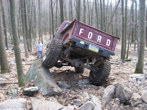 1991 Ford Bronco - A red Ford pickup truck is shown off-roading, with one tire on a large rock and the rear end angled upwards.