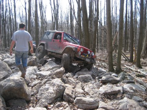 1991 Ford Bronco - A red Jeep Wrangler navigates a rocky trail in a forest, with a person walking nearby.