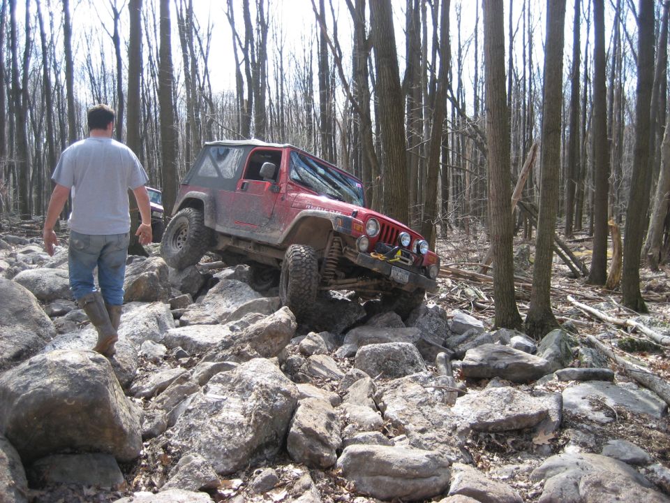 Red Jeep Wrangler climbing over rocks in a wooded area. Another vehicle is visible behind it