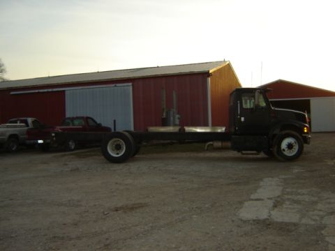 1992 Ford Bronco - A black truck chassis sits idle on a gravel lot next to a large red barn.
