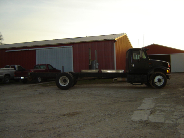 Black truck chassis with dual rear wheels parked on gravel in front of a red barn