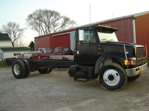 1992 Ford Bronco - A black cab-over truck chassis is parked in front of a red barn, with two pickup trucks visible in the background.