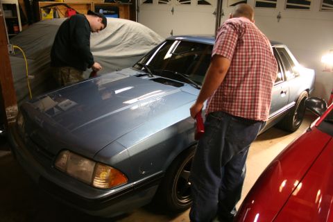 1990 Ford Mustang - Two people work on a blue 1990 Ford Mustang in a garage, with a red car partially visible.