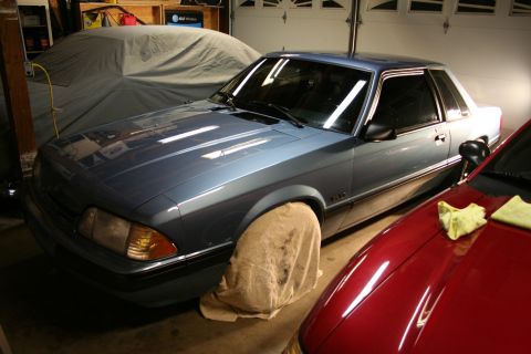 1990 Ford Mustang - A blue 1990 Ford Mustang coupe is parked in a garage next to a red car, with its front wheel covered.