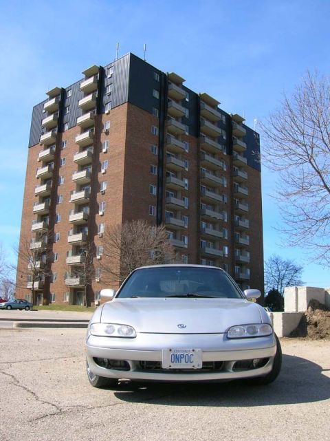 1994 Mazda MX-6 - A silver coupe is parked in front of a tall brick apartment building on a sunny day.