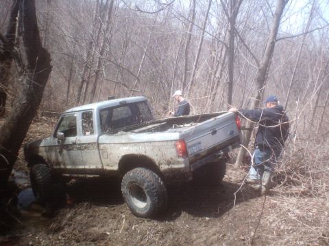 1990 Ford Ranger - A Ford pickup truck is stuck in the mud in a wooded area, with two people attempting to push it free.